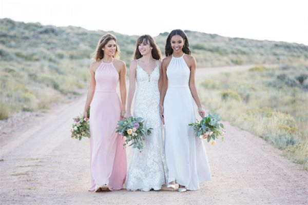 three women walking on a dirt road in bridesmaid dresses holding bouquet of flowers